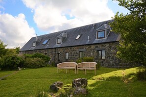 A stone building with a sloped roof, two windows, and a grassy lawn with two wooden benches.