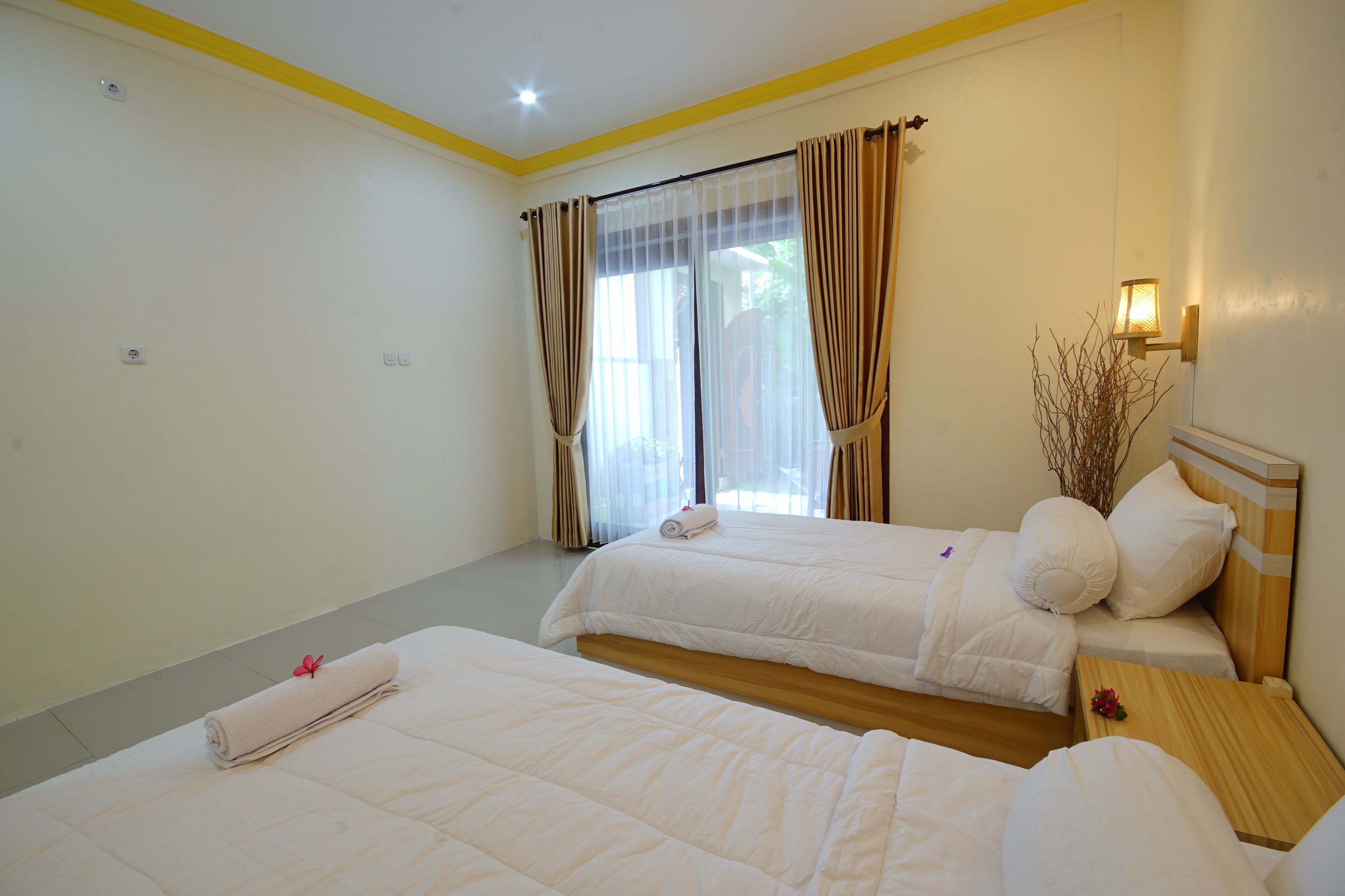 A hotel room with two beds, a wooden headboard, a nightstand, a lamp, and a window with curtains.