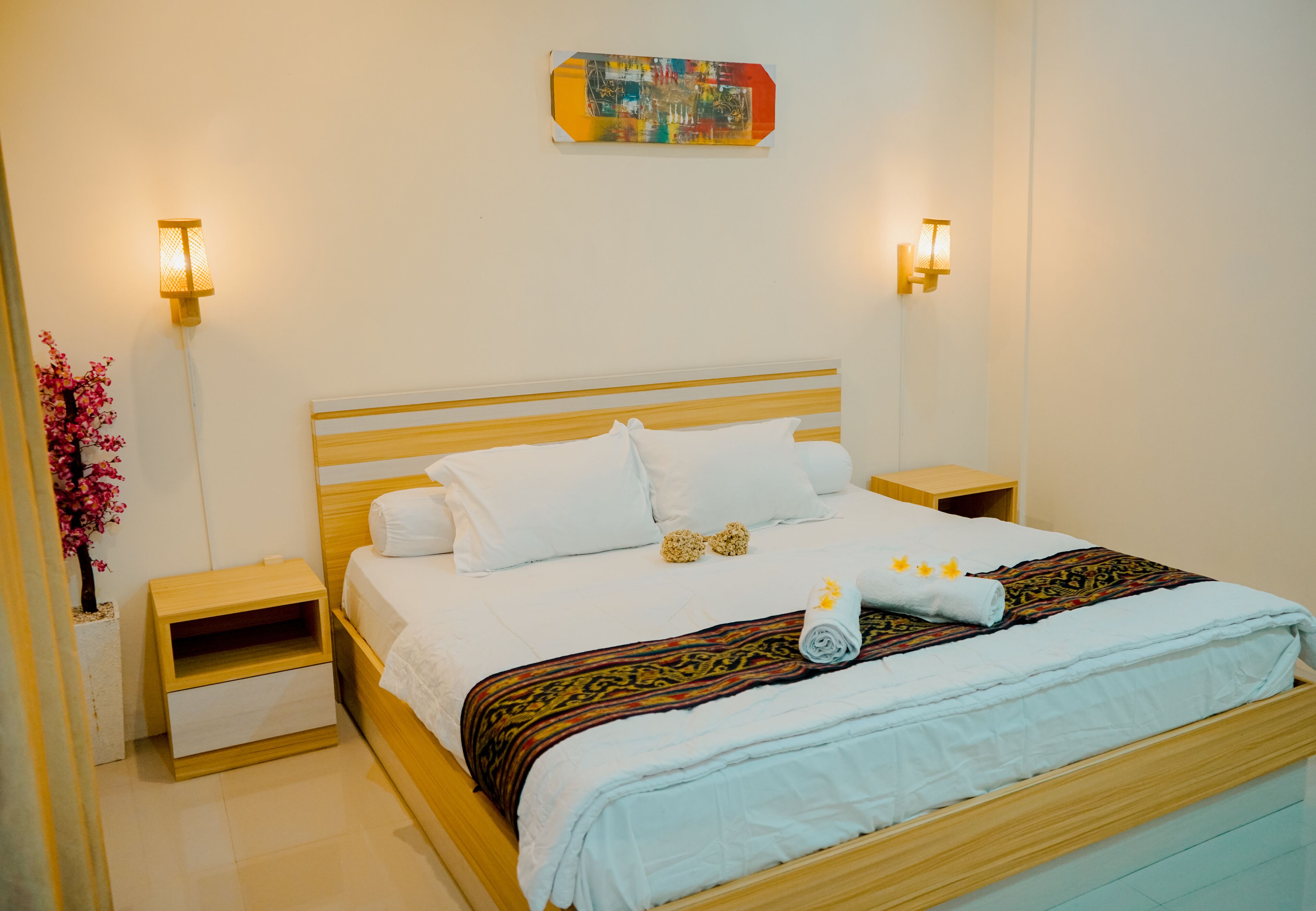 Uma cama bem arrumada com lençóis brancos e um cobertor estampado, duas mesas de cabeceira com luminárias e uma obra de arte pendurada na parede acima da cama.