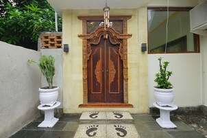 Uma porta dupla de madeira com detalhes em entalhe, ladeada por dois vasos de pedestal brancos com plantas em vasos.