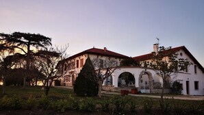 Un bâtiment à deux étages, doté d’un toit recouvert de tuiles rouges et de murs blancs, avec une porte d’entrée centrale en arc de cercle et une petite tour surmontée d’une cheminée.
