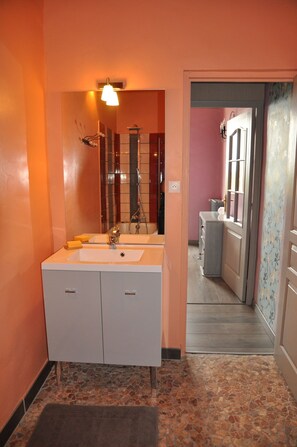 Une salle de bain avec deux lavabos, un miroir et une douche. Une porte donne accès à une autre pièce.