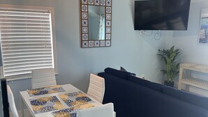 A modern living room with a dining area, featuring a wooden ceiling, a TV mounted on the wall, and a large mirror with a decorative frame. - Matlacha Sunset Cottage (Matlacha)