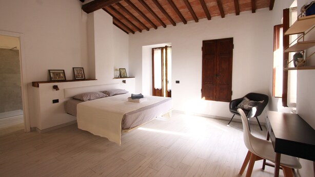A bedroom with a bed, a desk, and a chair. There is a wooden ceiling and a window.
