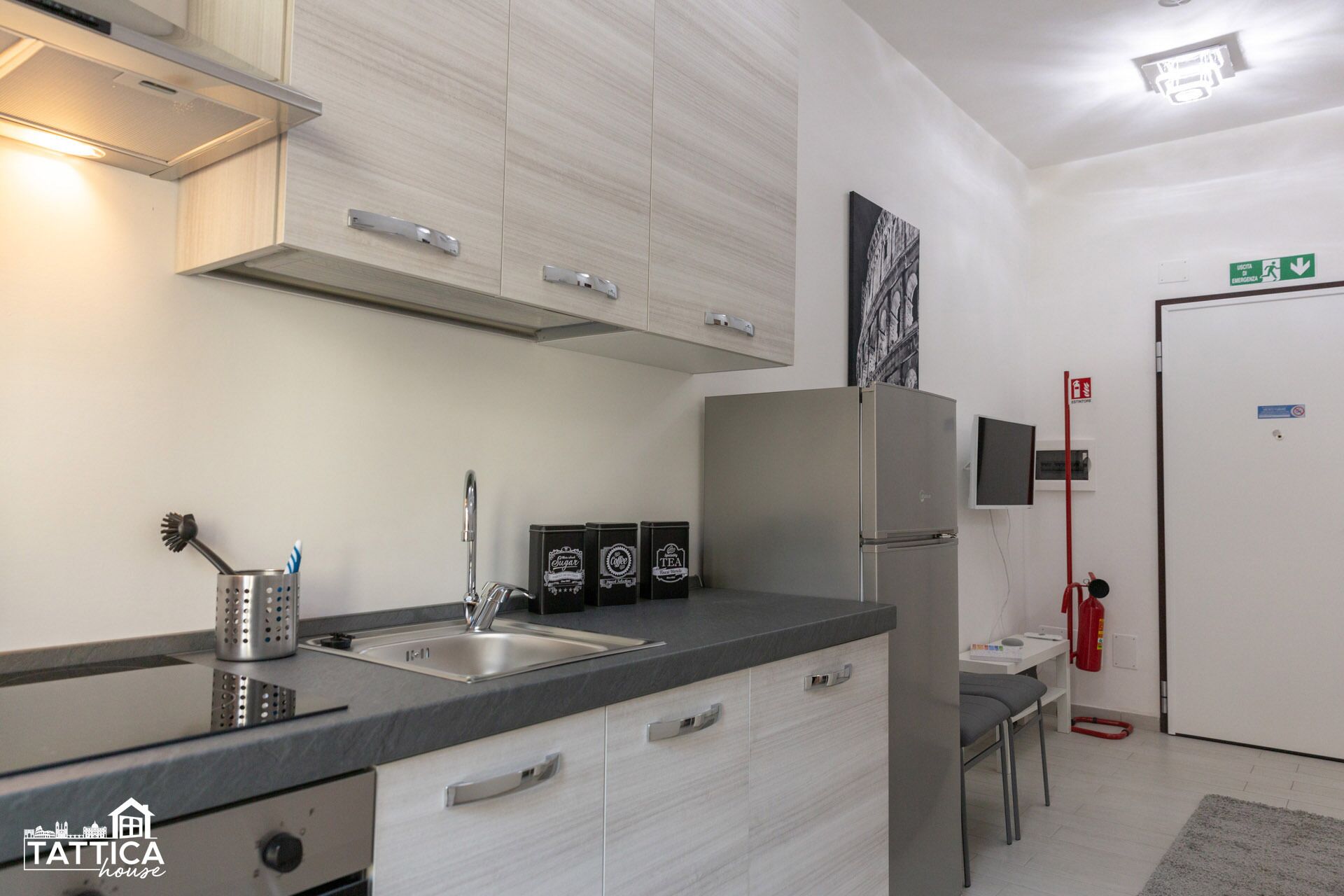 Une cuisine moderne équipée de deux éviers, d’un réfrigérateur en acier inoxydable et de placards dotés de poignées argentées. Un extincteur est fixé au mur et un tuyau d’incendie est posé sur le sol.