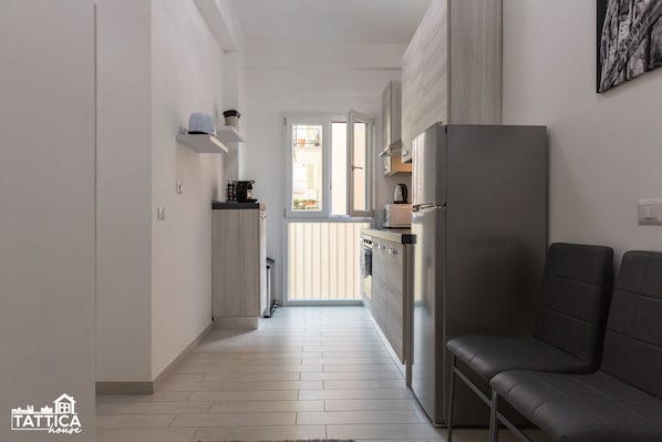A narrow hallway with a kitchen area on one side, featuring a stainless steel refrigerator, a microwave, and a stove. There is a window providing natural light, and a black leather sofa is visible on the right.