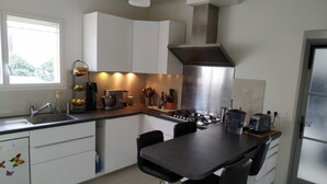 Una cocina moderna con armarios blancos, estufa a gas, cafetera y un comedor con dos taburetes tipo barra.