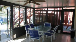 A sunlit patio with a glass table and chairs, a ceiling fan, and a view of a wooden deck and palm trees.