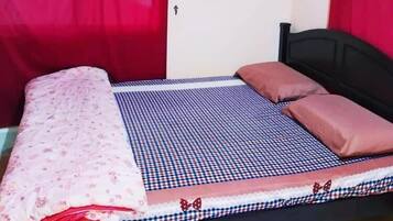 Un lit simple avec une couvre-lit à carreaux, une couverture rose et deux oreillers. Il y a un rideau rouge d’un côté et un mur blanc avec une porte.