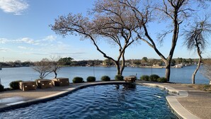 A clear swimming pool with a curved edge, surrounded by a pebble-lined deck and two wicker lounge chairs. In the background, there is a lake with boats and a tree-lined shore. - Lakefront property with pool and lighthouse with amazing views of the lake, . (Granbury)