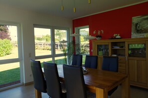 Un coin repas avec une table et des chaises en bois, de grandes fenêtres et un mur rouge orné d’un tableau.