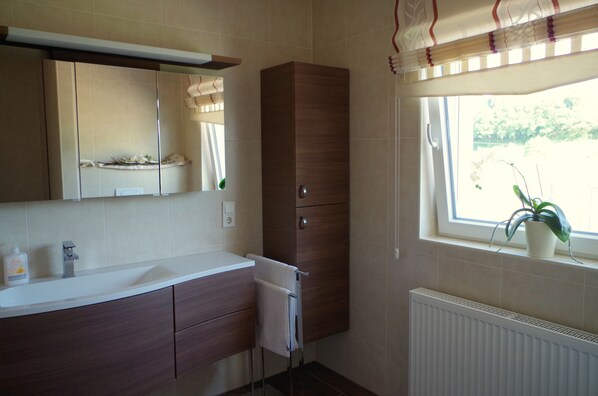 Une salle de bain avec un lavabo blanc, des meubles en bois, un porte-serviettes, un miroir et une fenêtre avec une plante.