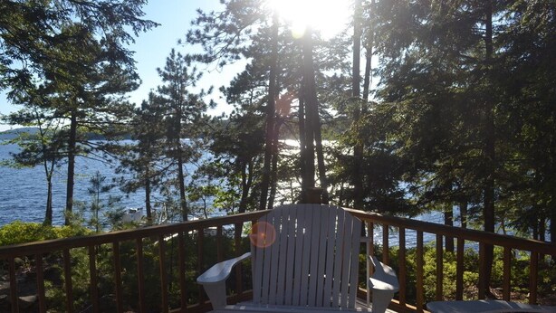 A white Adirondack chair on a wooden deck with a railing, overlooking a body of water and a forested area. - 10+ Spacious Cape on Winnipesaukee Island. Private Peninsula. Sunset Views. (Meredith)
