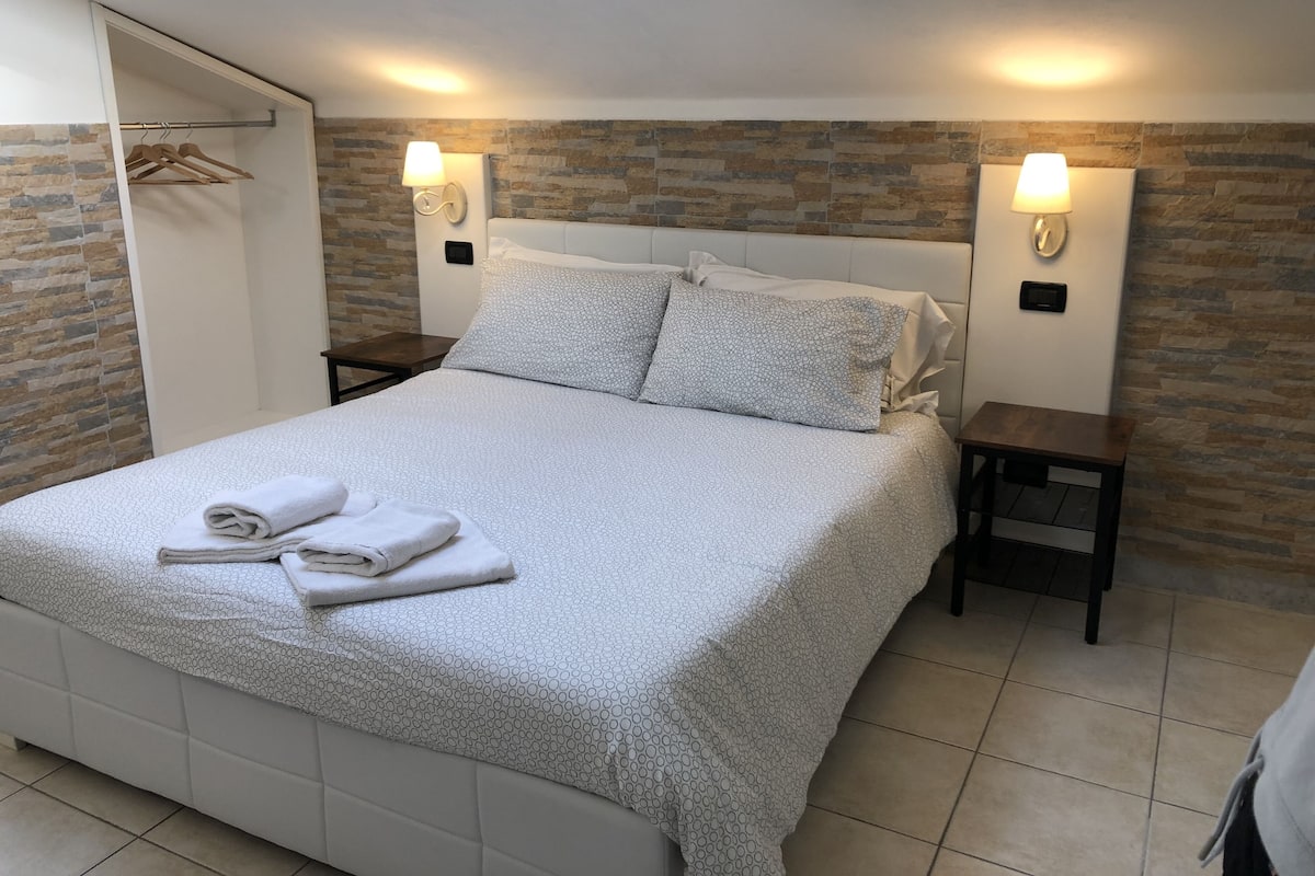 A neatly made bed with a patterned bedspread and pillows, two bedside tables with lamps, a wooden wardrobe, and a stone-patterned wall.
