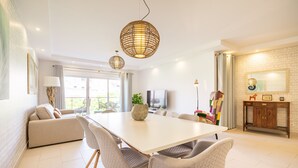 A modern living room with a white dining table, grey upholstered chairs, a sofa, and a TV. There are two hanging wicker lamps, a cabinet with decorative items, and a large window with curtains. - VERASL T - Albufeira - New, Luxury & comfy 2 bedrooms apartment (Albufeira)
