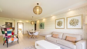 A modern living room with a sofa, coffee table, and a colorful armchair. There are two pendant lights, a dining table with chairs, and framed artwork on the wall. - VERASL T - Albufeira - New, Luxury & comfy 2 bedrooms apartment (Albufeira)