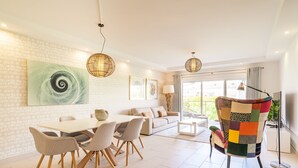 A modern living room with a dining area, featuring a white brick wall, a large abstract painting, a sofa, a colorful armchair, and a dining table with chairs. - VERASL T - Albufeira - New, Luxury & comfy 2 bedrooms apartment (Albufeira)