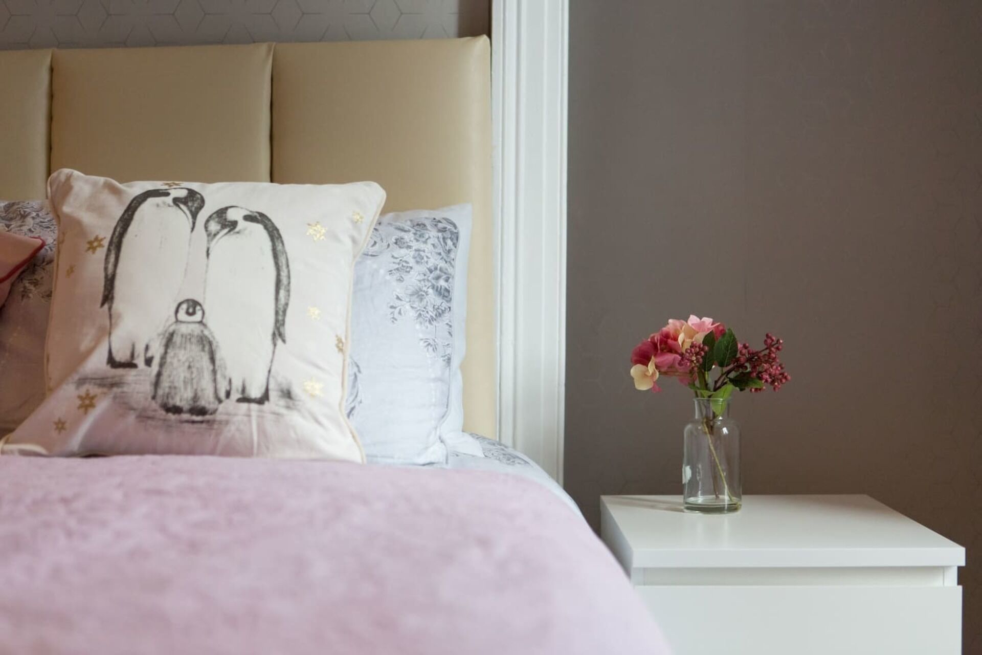 A neatly made bed with a pink blanket, a pillow with a penguin design, and a vase with flowers on a bedside table.