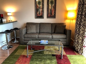 A living room with a grey sofa, two black bar stools, a glass coffee table, and a patterned rug.