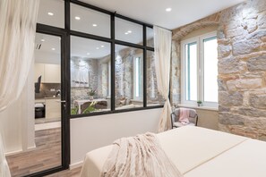 A bedroom with a stone wall, a large window, and a view of the kitchen.