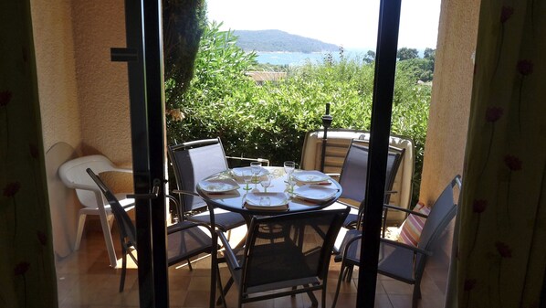 A balcony with a table set for four, surrounded by chairs, overlooking a lush garden and a distant view of the sea.