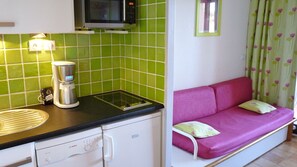 A kitchenette with a coffee maker, microwave, and a small refrigerator. There is a pink sofa with cushions and a green curtain with a floral pattern.