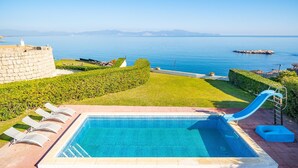 Una piscina con tobogán, rodeada de un patio de ladrillo y una zona de césped con vistas al mar.