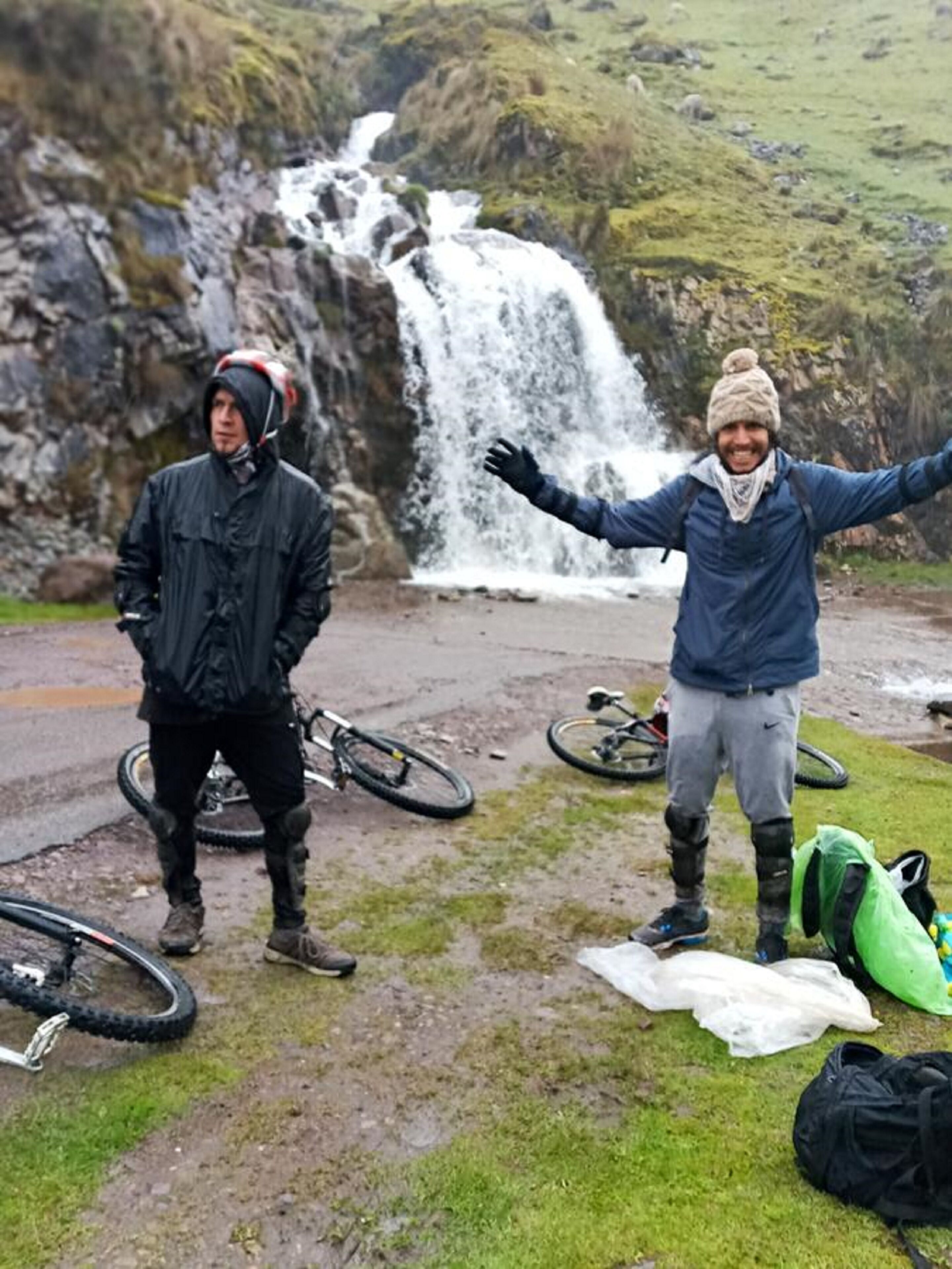 Twee personen staan voor een waterval, met fietsen en uitrustingstassen op de grond.