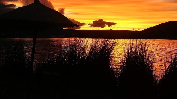 A sunset over a body of water with silhouettes of hills and a structure on the left. - Uros Titicaca Marca Lodge (Puno)
