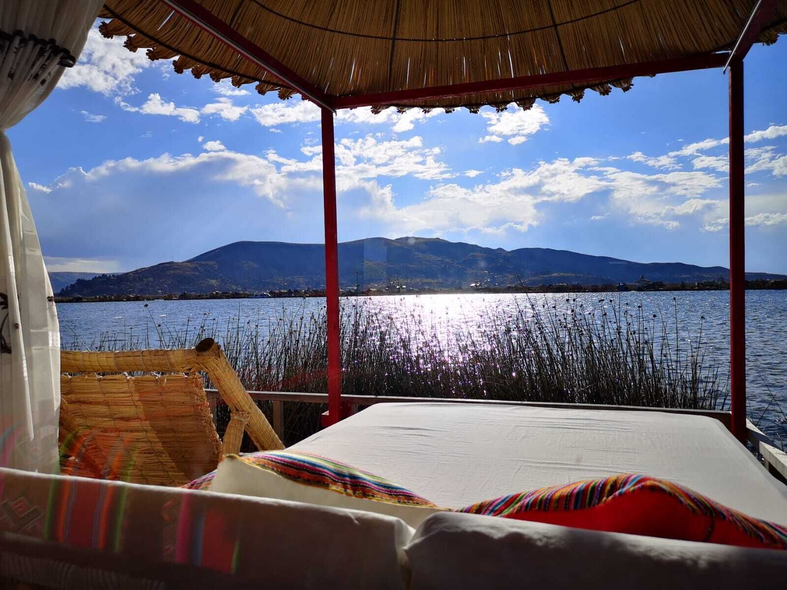 A boat with a thatched roof and a bed with colorful pillows, overlooking a calm lake with mountains in the background.