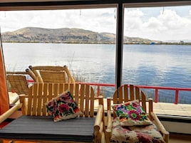 A wooden deck with two wooden chairs and cushions, overlooking a calm lake with distant hills.