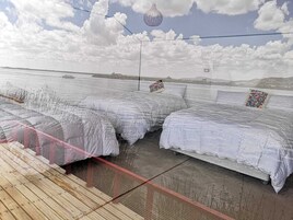 Un espacio amplio y abierto con filas de camas cubiertas, una terraza de madera y vistas a un cuerpo de agua y colinas distantes.