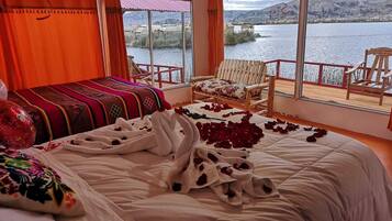 A bedroom with a bed, a chair, a window with a view of a lake and mountains, and a balcony with chairs.