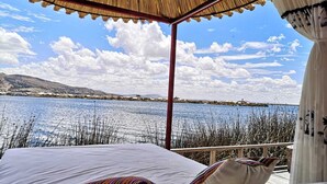A bed with white linens and colorful pillows, a thatched roof, and a view of a lake with reeds and distant buildings. - Uros Titicaca Marca Lodge (Puno)