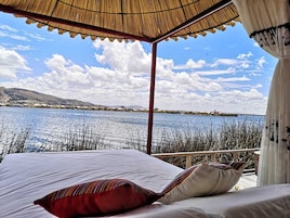 A bed with white linens and colorful pillows, a thatched roof, and a view of a lake with reeds and distant buildings.