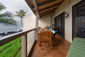 A balcony with a wooden table and chairs, a view of the ocean, and a tropical setting with palm trees.