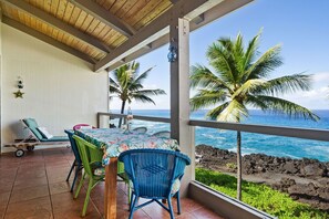 A covered outdoor seating area with a table and chairs, overlooking the ocean with palm trees and a rocky shoreline.