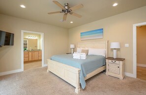 Ein Schlafzimmer mit Bett, Nachttischen, Deckenventilator, einem Gemälde und einem durch eine offene Tür einsehbarer Badezimmer.
