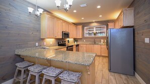 Cocina moderna con armarios de madera, encimeras de granito, refrigerador de acero inoxidable y una barra con taburetes.