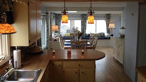 Een keuken met houten kasten, een centraal eiland, hanglampen, en een eethoek met een tafel en stoelen.