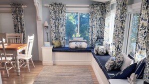 Een gezellige woonkamer met een houten vloer, een ingebouwde bank met kussens, een eettafel met stoelen en een groot raam met bloemen gordijnen.