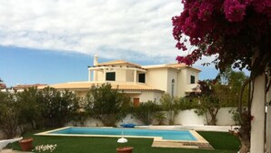 A two-story house with a swimming pool, surrounded by greenery and a clear sky.