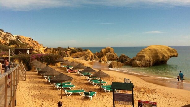 A beach with green lounge chairs and umbrellas, rocky formations, and a wooden walkway.