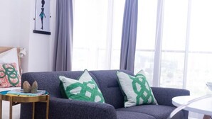 A modern living room with a dark grey sofa, two patterned throw pillows, a small side table with a book and a cup, and a view of the city through large windows with grey curtains.