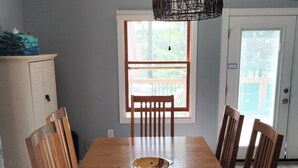 Une salle à manger avec une table en bois et six chaises, un luminaire suspendu, une fenêtre donnant sur un paysage et une porte.