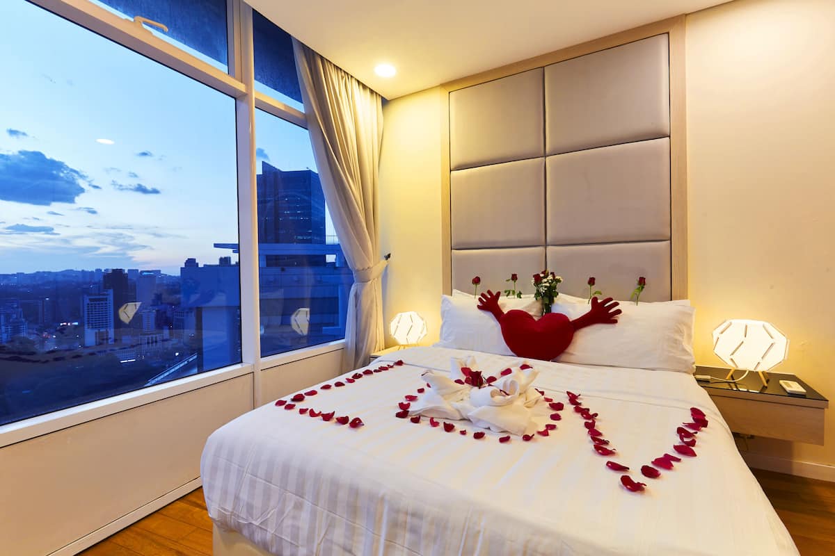 Ein Hotelzimmer mit großem Fenster, einem Bett mit Rosenblättern, einem herzförmigen Kopfkissen und Blick auf die Stadt.