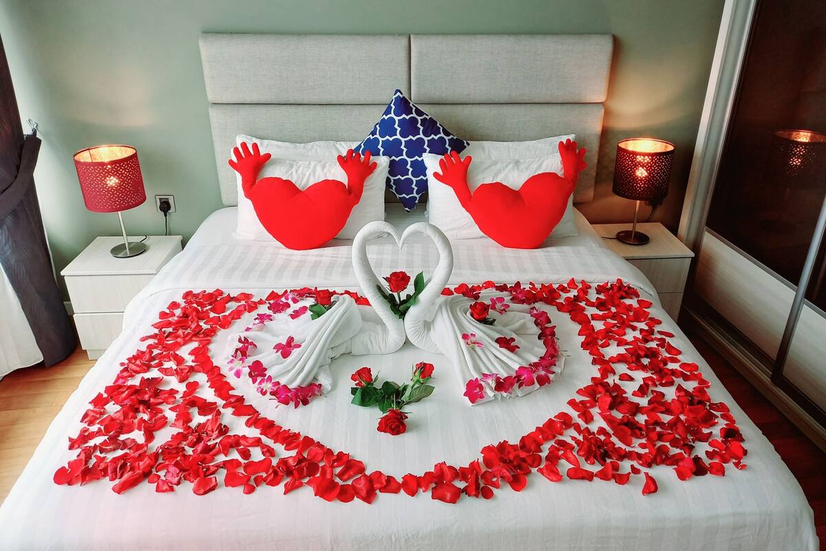 Ein ordentlich bezogenes Bett mit roten Rosenblättern und einer Schwan-Figur, zwei herzförmigen Kissen und ein blaues Kissen mit Herzmuster.