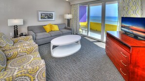 A hotel room with a grey sofa, yellow pillows, a white coffee table, a red dresser, and a television. There is a ceiling fan and a window with a view of the ocean.