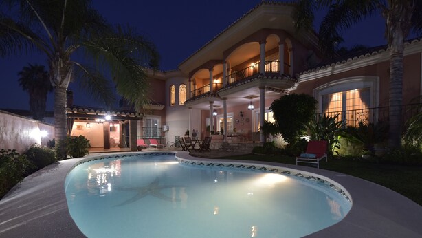 Une piscine entourée d’une maison dotée d’un balcon, de palmiers et de mobilier d’extérieur.