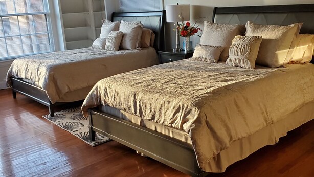 A bedroom with two beds, wooden flooring, a window with blinds, and a bedside table with a lamp and vase. - Ida's Oasis  near JRMC Hospital ,  Pine Bluff Country Club & Restaurants . (Pine Bluff)